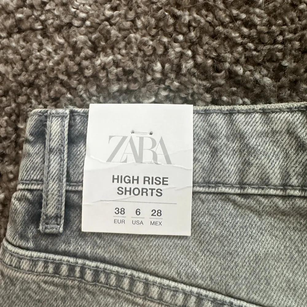 Ett par grå jeansshorts från Zara med hög midja. Shortsen har en rå kant och slitna detaljer vid fickorna. De har en knapp och dragkedja framtill samt fem fickor.. Shorts.