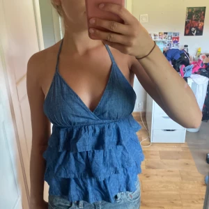 Blå jeanslinnetopp med volanger - Säljer en superfin blå jeanslinnetopp med volanger. Toppen har smala axelband som knyts i nacken och en öppen rygg. Perfekt för sommaren och kan stylas både till vardags och fest. Toppen är i bra skick och väldigt bekväm att ha på sig. Passar perfekt till ett par jeansshorts eller en kjol!