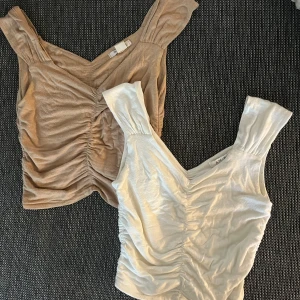 Två ärmlösa toppar i beige och vit - Säljer två snygga ärmlösa toppar i beige och vit. Båda topparna har en rynkad design framtill och breda axelband. Perfekta för sommaren och kan stylas både till vardags och fest. Materialet känns mjukt och bekvämt mot huden.