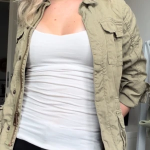Grön jacka med text på ryggen - Säljer en beige jacka med texten 'IS IT FRIDAY YET?' på ryggen. Jackan har en dragkedja framtill och långa ärmar. Den är perfekt för vår och höst och har en avslappnad, normal passform. Jackan har en cool casual stil. Finns en fläck på en av fickorna, skriv för fler bilder☺️ Lite kort i armarna 