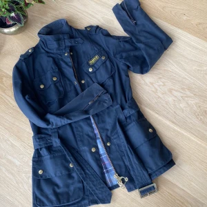 Svart jacka från Barbour - Säljer en snygg svart jacka från Barbour. Jackan har en klassisk design med flera fickor och en dragkedja framtill. Den har också ett bälte i midjan för en bättre passform. Insidan är fodrad med ett stilrent rutigt mönster. Perfekt för höst och vår!