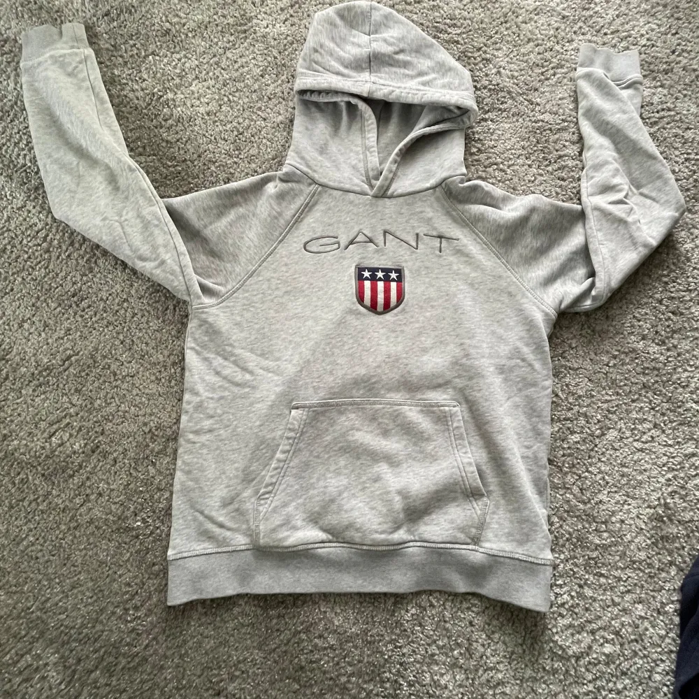 Säljer en snygg och bekväm grå hoodie från GANT. Den har en stor ficka på magen och ett coolt emblem med amerikanska flaggan på bröstet. Perfekt för höst och vinter, och den är i bra skick. Passar perfekt till vardags eller chillkvällar. 🧥. Hoodies.