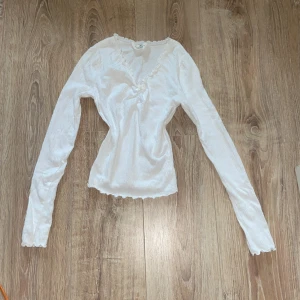 Vit långärmad tröja från H&M - Säljer en söt vit långärmad tröja från H&M. Den är i storlek 158/164 i barnstorlek men passar även någon i SX. Den har en v-ringad hals och små spetsdetaljer runt halsen och ärmsluten. Tröjan är i ett mjukt och bekvämt material.