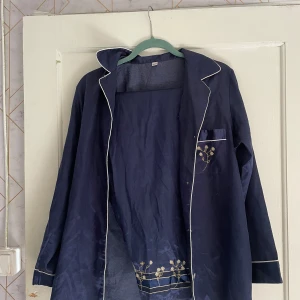 Mörkblå långärmad jacka med blommigt broderi - Säljer en mörkblå långärmad pyjamas sett med vita detaljer och ett fint blommigt broderi på fickan och nederkanten. Jackan har en avslappnad passform och är perfekt när man ska sova. Den är i storlek 38/40 och i bra skick. Perfekt när man sover då den inte är så varm. 
