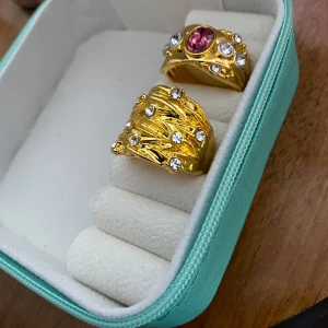 Två guldpläterade ringar med stenar - Säljer två snygga guldpläterade ringar. Den ena ringen har en stor rosa sten i mitten och flera små klara stenar runtom. Den andra ringen har en flätad design med små klara stenar utspridda. Perfekta för att ge en touch av glamour till din outfit! 💍✨