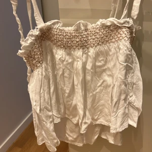 Vit ärmlös topp med smockdetaljer - Säljer en söt vit ärmlös topp med smockdetaljer runt bysten och volangkant. Toppen har tunna axelband och är perfekt för sommaren. Materialet är lätt och luftigt, vilket gör den bekväm att bära under varma dagar. Passar både till vardags och festliga tillfällen. Aldrig använts! Köptes för 499kr