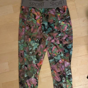Färgglada leggings med blommönster - Säljer ett par färgglada leggings med ett coolt blommönster. De har en bred resår i midjan med texten 'SPORT' och är perfekta för träning eller en avslappnad dag. Materialet känns som syntet och de är superbekväma att ha på sig. Passar perfekt för dig som vill sticka ut lite extra!