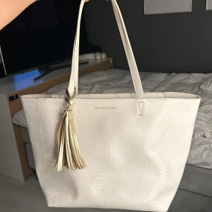 Vit axelväska från Victoria's Secret - Säljer en snygg vit axelväska från Victoria's Secret. Väskan har en stilren design med ett mönster som liknar ormskinn. Den har en praktisk dragkedja upptill och en dekorativ tofs i guld. passar bra till skolan!