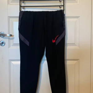 Svarta träningsbyxor från Nike - Säljer ett par svarta träningsbyxor från Nike i storlek L. Byxorna är slim fit och har en röd Nike-logga på sidan. De är tillverkade i ett Dri-FIT material som håller dig torr och bekväm under träning. Byxorna har elastisk midja med snörning och fickor på sidorna.