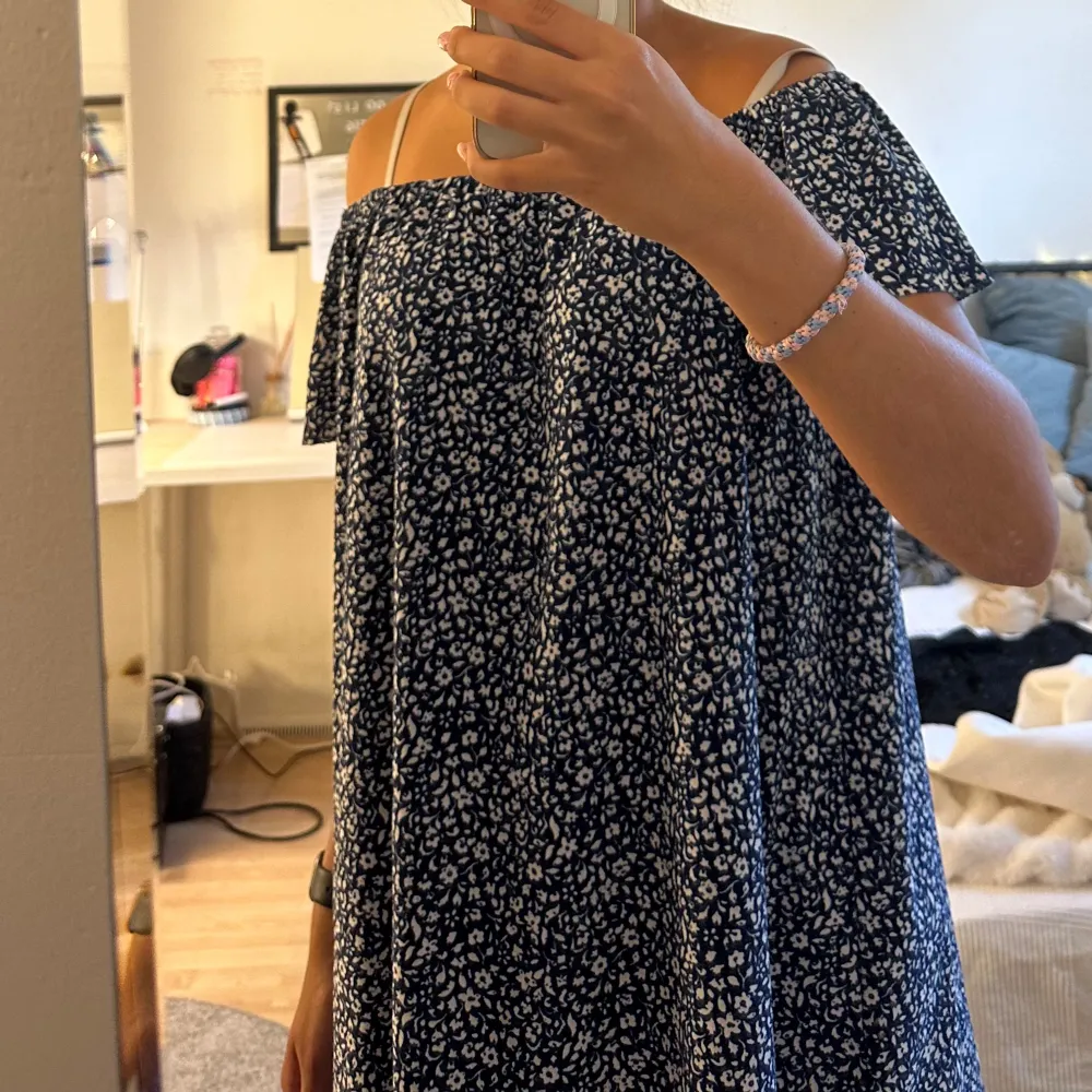 Säljer en superfin blommig offshoulder klänning. Den är marinblå med vita små blommor över hela tyget. Klänningen är kort och har en lös passform, perfekt för sommaren eller en fest.. Den har korta ärmar och en söt volangdetalj vid axlarna. Perfekt för en avslappnad dag eller en somrig fest!. Klänningar.