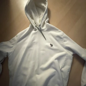 Vit hoodie från Paul Smith - Säljer en snygg och bekväm krämvit hoodie från Paul Smith. Den är i storlek L och har en klassisk design med en liten logga på bröstet. Perfekt för både vardag och avslappnade tillfällen. Hoodien är i bra skick och väldigt mjuk och skön att ha på sig. Passa på att köpa den här stilrena hoodien!