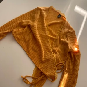 Gul ribbad omlottopp - En gul ribbad omlottopp med långa ärmar och knytband i midjan. Toppen har en v-ringad design och är tillverkad i ett mjukt och bekvämt material.