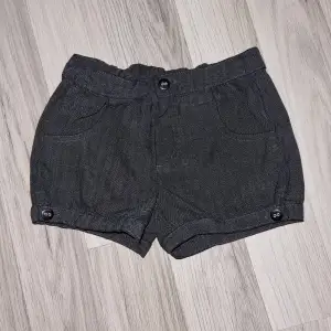 Ett par mörkgrå shorts med knappar framtill och vid bensluten. Shortsen har två framfickor och en elastisk midja. Från Lindex i storlek 110/116