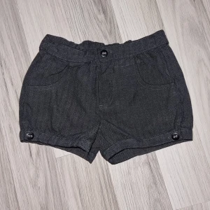 Grå shorts med knappar - Ett par mörkgrå shorts med knappar framtill och vid bensluten. Shortsen har två framfickor och en elastisk midja. Från Lindex i storlek 110/116