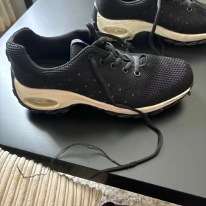 Svarta sneakers med lila detaljer - Ett par svarta sneakers med lila detaljer och snörning. Skorna har en vit sula och texten 'FASHION' på plösen.