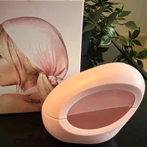 Mod Blush Parfym - En elegant parfym från Ariana Grande med namnet Mod Blush. Flaskan är oval och ljusrosa, och förpackningen har en bild av en kvinna med en rosa sjal på huvudet.