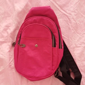 Rosa axelväska - En praktisk och stilren rosa axelväska med flera fack och dragkedjor. Väskan har en justerbar svart axelrem och en knappdetalj på framsidan.