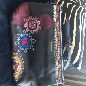 Svart axelväska från Desigual - En svart axelväska från Desigual med färgglada broderade mönster. Väskan har en justerbar axelrem och flera fack med dragkedja. Insidan är fodrad med blått tyg och har ytterligare fack för förvaring.