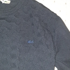 Mörkblå tröja från Lacoste - Utan defekter. Står L men passar som  M Mörkblå/ svart tröja från Lacoste med ett diskret mönster och Lacoste-logotypen broderad på bröstet. 