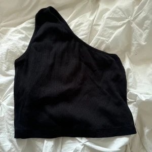 Svart oneshoulder topp - En svart oneshoulder topp i ribbat material. Toppen är ärmlös och har en asymmetrisk design med en axelbandslös sida.