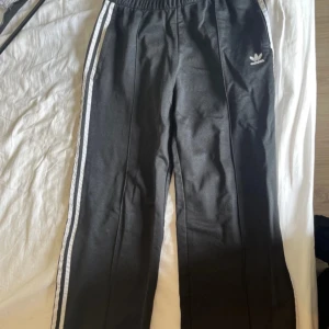 Svarta träningsbyxor från Adidas - Ett par svarta träningsbyxor från Adidas med vita ränder längs sidorna. Byxorna har en elastisk midja och Adidas-logotypen på vänster ben.