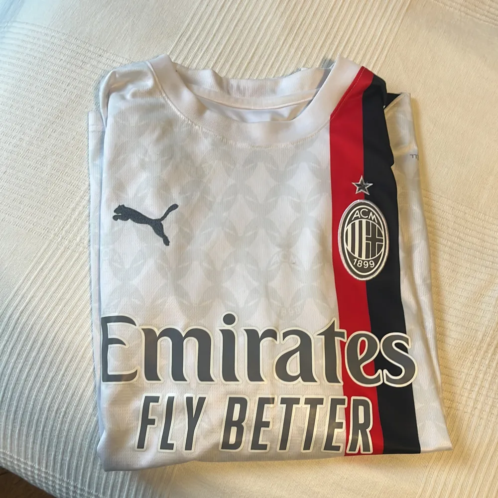 En vit AC Milan fotbollströja från Puma med klubbens emblem och sponsorer tryckta på framsidan. Tröjan har en röd och svart rand på vänster sida och är märkt med 'Emirates Fly Better' och 'wefox'. Ärmarna har Puma-logotypen och MSC-logotypen tryckta. Tröjan är i storlek M och är tillverkad i Thailand.. Sportswear.