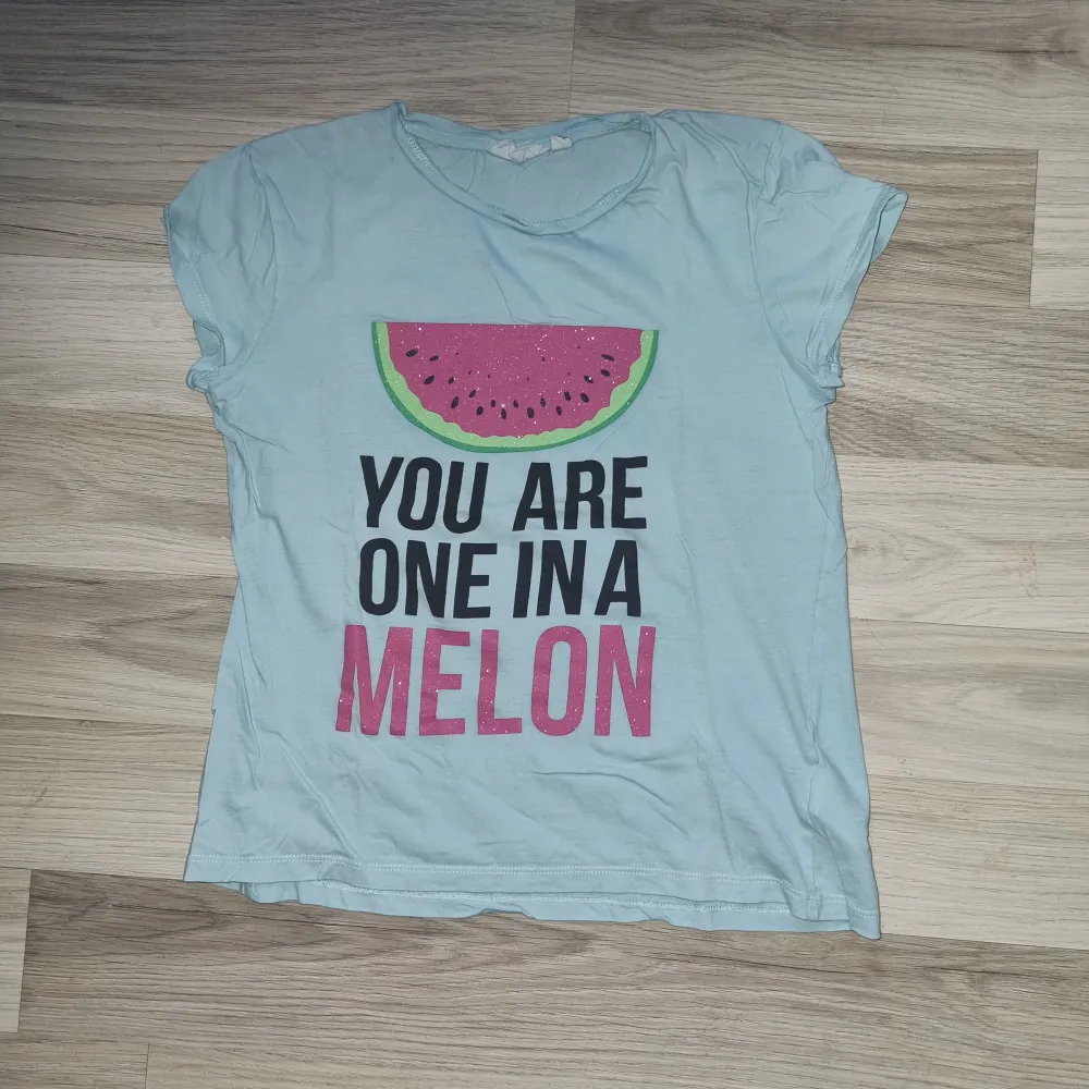 En ljusblå t-shirt med ett tryck av en vattenmelon och texten 'You are one in a melon' i svart och rosa. T-shirten har korta ärmar. Från kappahl i stl 146/152. T-shirts.