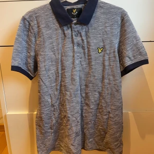 Lyle & Scott T-shirt - Oanvänd Gråblå pikétröja från Lyle & Scott. Inköpt på Lyle & Scott för ca 1år sedan. Normal i storleken. (Finns en extra knapp med).