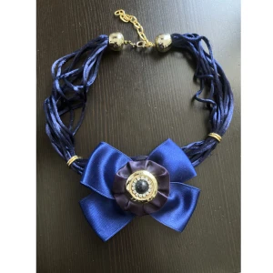 Mörkblått halsband med rosett, marinblå, silver, guld, satin, strass - Mörkblått halsband i satin, silverfärgade delat i plast och guldfärgade i metall, justerbar. Finns lite slitage på snörena. Längd: 46-54 cm, bredd rosett: 9,5 cm.