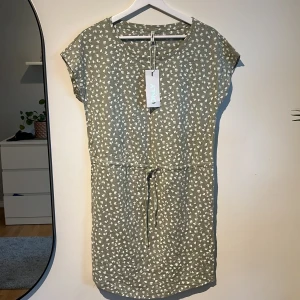 Olivgrön med vita blommor - Säljer en olivgrön klänning som är aldrig använd. Prislappen hänger kvar också. Köpt för 130kr och säljer för 100kr istället. Ett snöre vid midjan som du kan göra en rosett av. Gott skick! 