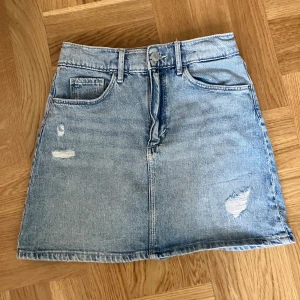 Kort ripped jeanskjol - Mått: 140 cm. Skick: Perfekt skick. 34 cm fram och 35 cm bak. Justerbar midja. 