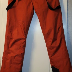 Neomondo skidbyxor från XXL - Vatten- och vindtäta. 10.000 pelare regntäthet. Justerbar midja, justerbara/avtagbara hängslen Från XXL.  Färgen är tegelfärgad/roströd.