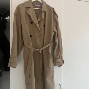 Lång trenchcoat  - Säljer min långa beiga/bruna trenchcoat från danska hemsidan BYIC. Den är i storlek M, jag är vanligtvis en S och 164 cm lång och denna sitter perfekt oversize på mig! Köptes hösten 2022 och är använd men har inga defekter, fläckar eller liknande. 
