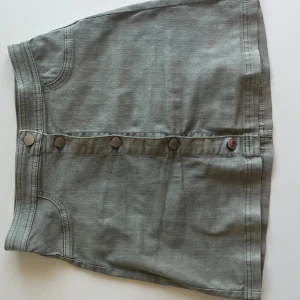 Grön/Grå Jeans kjol med silvriga knappar  - Jag säljer en militärgrön/gråaktig kjol från H&M. Kjolen är gammal från H&Ms barnavdelning och jag skulle säga att den passar en XS/S. Inga defekter och bra sick! 