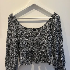 Svart blommig topp från BikBok - En svart topp från BikBok med ett vitt blommönster. Toppen har 3/4 ärmar och en elastisk kant vid axlarna för en bekväm passform. Den har också en volangdetalj vid nederkanten.