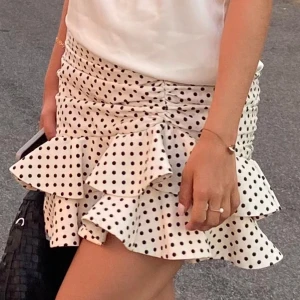 Vit kortkjol med svarta prickar - En vit kortkjol med svarta prickar och volangdetaljer. Kjolen har en tight passform och är dekorerad med flera lager av volanger.