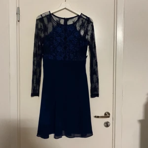 Mörkblå klänning med spetsdetaljer - En elegant mörkblå klänning med spetsdetaljer på överdelen och ärmarna. Klänningen har långa ärmar och en rund halsringning. Kjoldelen är enkel och faller fint.💕