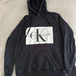 Svart hoodie från Calvin Klein Jeans - En svart hoodie från Calvin Klein Jeans med en stor vit logotyp på framsidan. Hoodien har en justerbar huva med dragsko och en stor ficka på magen. Den har långa ärmar med ribbstickade muddar vid ärmslut och nederkant. 100% Bomull