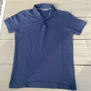 Mörkblå pikétröja från J.Lindeberg - En stilren mörkblå pikétröja från J.Lindeberg. Tröjan är tillverkad av 100% bomull och har korta ärmar samt en klassisk krage med knappar.