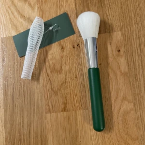 Grön sminkborste från Avon - En sminkborste med ett grönt handtag och vita borst från Avon. Borsten har ett silverfärgat fäste och kommer med ett skyddsnät för borsten.