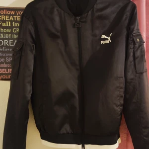 Svart bomberjacka från Puma - En svart bomberjacka från Puma med dragkedja framtill och vita Puma-logotyper på bröstet och ryggen. Jackan har långa ärmar och en ficka med knapp på ena ärmen.