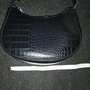 Svart axelväska  - Säljer denna eleganta svarta väska. Tillverkad av högkvalitativt material som efterliknar äkta läder. Den är generöst rymlig med måtten 22 cm i bredd och 15 cm i längd, med en bredd på 7 cm i botten. Endast använd några få gånger och i utmärkt skick.