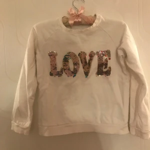 Vit tröja med paljetter  - Vit sweatshirt med paljetter i texten LOVE. Paljetterna går att vända åt två håll. Åt ena sidan är paljetterna silvriga och på andra sidan är de i olika färger. Tröjan är i ganska bra skick, det ända som jag hittar är en liten fläck (se sista bild).