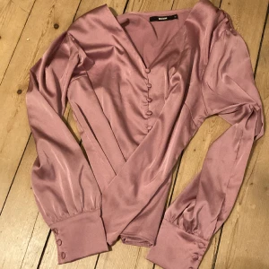 Rosa blus med vackra detaljer - Vacker rosa blus i sidenefterlikning. V-utrigning med tygklädda knappar ner långs mitten. Tygklädda knappar även vid handlederna. Kan passa 36-42