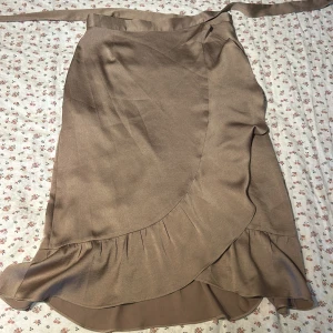 Beige omlottkjol från Neo Noir - En elegant beige omlottkjol från Neo Noir. Kjolen har en volangdetalj längs nederkanten och knytband i midjan. Den är tillverkad i ett mjukt och lätt material som ger en fin passform. Aldrig använd, nypris 699kr.