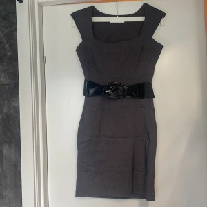 Mörkgrå klänning med bälte - En elegant mörkgrå klänning med en figurnära passform. Klänningen har breda axelband och ett svart bälte i midjan med en stor spänne. Den har en dragkedja på sidan för enkel på- och avklädning. bälte går att byta ut. lite defekt på bältet se bild fem. 