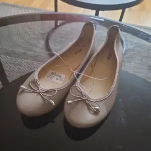 Ett par eleganta beige/bruna ballerinaskor med en dekorativ rosett på tån. Skorna har en rund tå och är tillverkade i ett syntetiskt material.