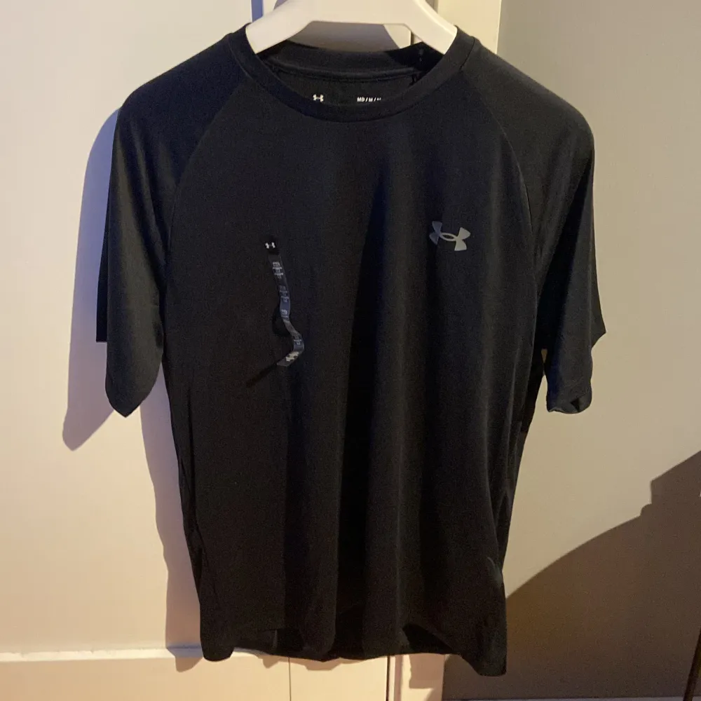 En svart t-shirt från Under Armour med korta ärmar och rund hals. T-shirten har en liten logotyp på bröstet och en större logotyp på ryggen. Den är tillverkad i ett lätt och andningsbart material, perfekt för träning.. T-shirts.