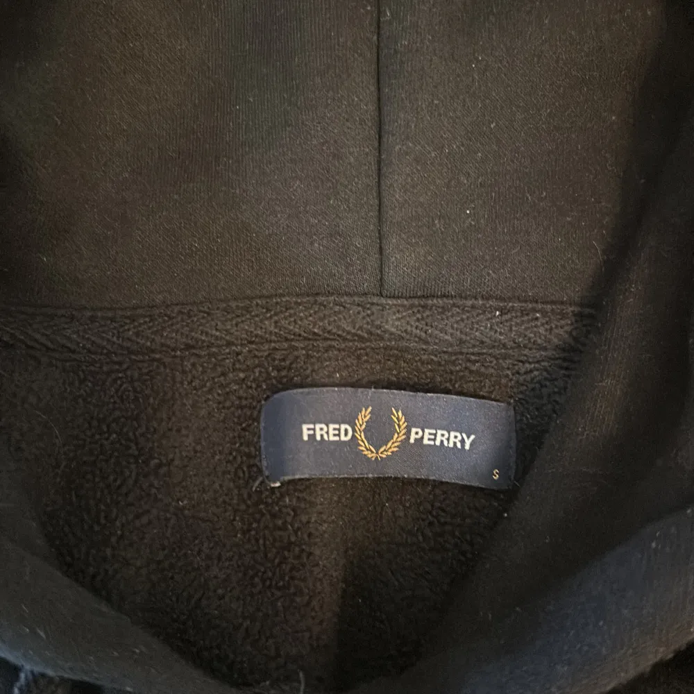 En svart hoodie från Fred Perry med en broderad logotyp på bröstet. Hoodien har långa ärmar och en justerbar huva med dragsko.. Tröjor & Koftor.