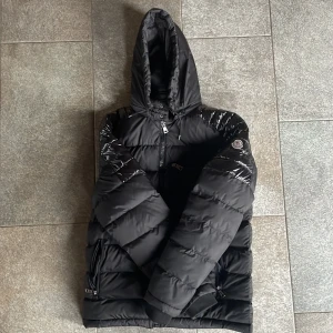 Moncler Jacka - En svart Moncler jacka med glansiga axlar. Detta är en äldre modell av moncler som inte görs längre.